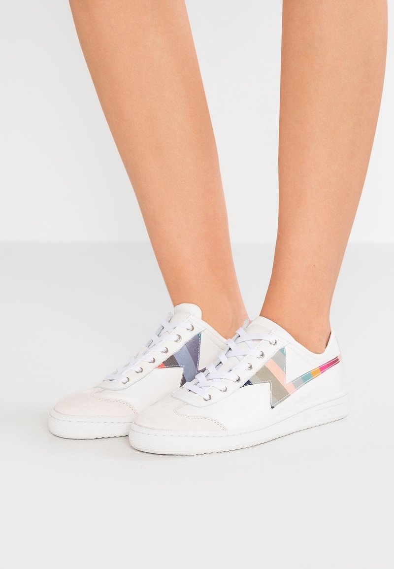 Baskets blanches basses avec des motifs géométriques colorés sur les côtés, portées pieds nus, debout sur un sol blanc uni.