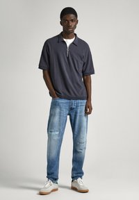 Polo de manga corta gris oscuro con cremallera en el cuello, combinado con unos vaqueros desgastados de color azul claro y zapatillas blancas.