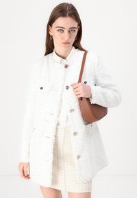 White tweed jacket with fringed edges, gold-tone buttons, and two front pockets, paired with a beige tweed skirt and a brown shoulder bag.
