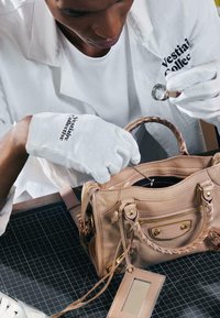 Personne portant des gants blancs et une blouse de laboratoire inspecte un sac à main en cuir beige à l’aide d’une loupe et d’un outil en métal.
