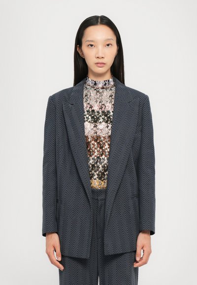Blazer en chevrons gris foncé avec une coupe ajustée et des revers en biais, porté sur un haut à motifs multicolores. Texture lisse, design structuré.