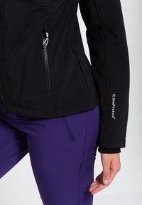 Gros plan sur une personne portant une veste noire zippée avec "clima protect" sur la manche et un pantalon violet avec une poche zippée.