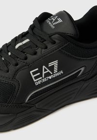 Zwarte sport sneaker met een gestructureerde bovenkant, mesh details en reflecterende accenten. Logo prominent aan de zijkant weergegeven.