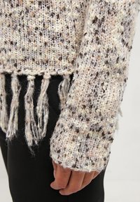 Pull tricoté avec un motif texturé en nuances de crème, beige et marron ; présente des détails en franges à l'ourlet et des manches longues.