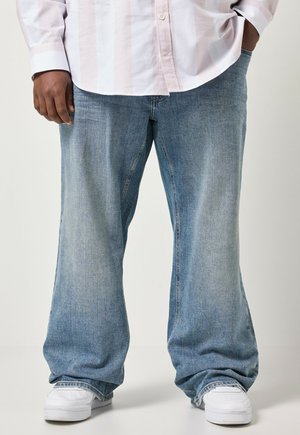 Persoon met lichtblauwe wijde jeans, witte sneakers en een licht gestreept overhemd, staand met één hand in de zak.