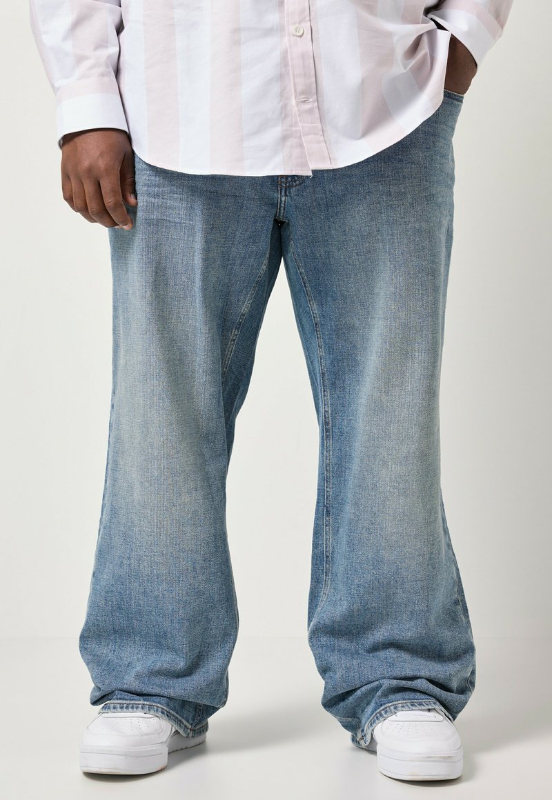 Person iført lyseblå vide jeans, hvide sneakers og en lyseblå skjorte med striber, stående med den ene hånd i lommen.