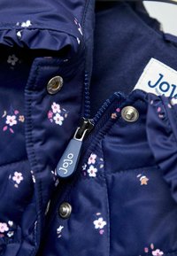 Mornarska dikcija jakna s majhnim cvetličnim vzorcem, modri zadrga z oznako "Jojo", kovinske zapiralo s pritiskom in nabran ovratnik za dodano teksturo.