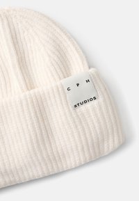 Valge ribitud kudumisega beanie, millel on pehme tekstuur ja õmmeldud silt, kus on mustade suurte tähtedega kirjas "CPH STUDIOS" valgel taustal.