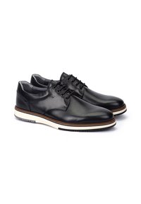 Par de zapatos de vestir negros de cuero con cordones y suelas de goma blancas y marrones sobre un fondo blanco.