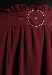 Dunkelrotes Stoffkleidungsstück mit geraffter Taille und breitem Gürtel, der mit einer großen silbernen runden Schnalle und Metallösen verziert ist.
