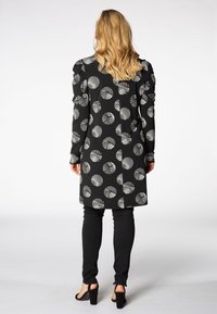 Manteau noir avec des motifs circulaires rayés blancs, manches longues, détail froncé à l'épaule, associé à un pantalon skinny noir et des talons.