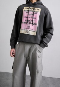 Felpa grigia antracite con stampa grafica in testo rosa e nero. Abbinata a pantaloni cargo grigi medi su misura con tasche laterali.