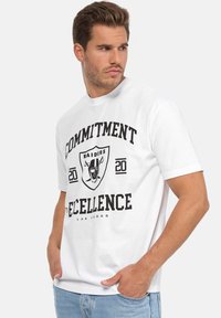 Camiseta de algodón blanca con texto impreso en negro y un gráfico de un escudo con la cara de un futbolista, con mangas cortas y cuello redondo.