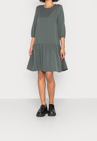 Vestido gris hasta la rodilla con mangas de tres cuartos, cintura fruncida y falda de capas. Combinado con zapatos de plataforma negros sin cordones.