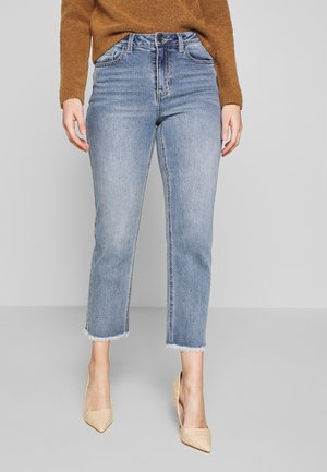 Frau trägt hellblaue, kurze Jeans mit ausgefransten Säumen, einen braunen Pullover und beige spitze High Heels, steht vor einem schlichten grauen Hintergrund.