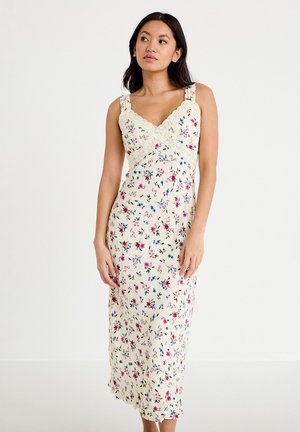 Kobieta ubrana w kremową, bezrękawnikową sukienkę maxi z wielokolorowym kwiatowym nadrukiem i koronkowym wykończeniem, stojąca na tle jednolitego białego tła.