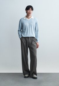 Mladý muž v světlomodrém svetru s bílým límečkem, tmavých širokých kalhotách, teniskách a perlovém náhrdelníku, stojící s rukama v kapsách.