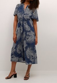 Žena v námořnicko-modrých midi šatech s bílým paisley vzorem a volnými rukávy, doplněná černými sandály na blokovém podpatku se svršky.