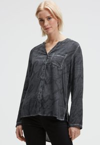 Blonde Frau, die eine dunkelgraue Bluse mit dezentem Schriftzug und schwarze Hose trägt, steht vor einem einfarbigen hellen Hintergrund.