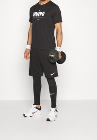 Camiseta negra de Nike con el texto "HWPO", pantalones cortos negros y mallas compresoras negras. Sosteniendo un dumbbell negro. Zapatillas blancas de Nike.