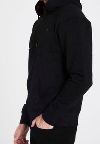 Sudadera negra de tela suave, con un bolsillo frontal, capucha con cordón y un pequeño logo en el pecho. Acento rojo en el interior de la capucha.