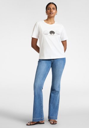 Kobieta ubrana w białą koszulkę z trzema czarno-białymi nadrukami muszli i niebieskie dżinsy, stoi z rękami za plecami.