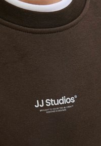Brūna kokvilnas sporta jaka ar ribotu apkakli. Ir balts teksts: "JJ Studios piedāvā J&J komanda. Izstrādāts Dānijā."