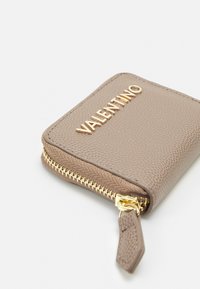 Portafoglio in pelle marrone con texture, logo "VALENTINO" in tonalità oro, chiusura a zip e una piccola linguetta in pelle. Design compatto e rettangolare.