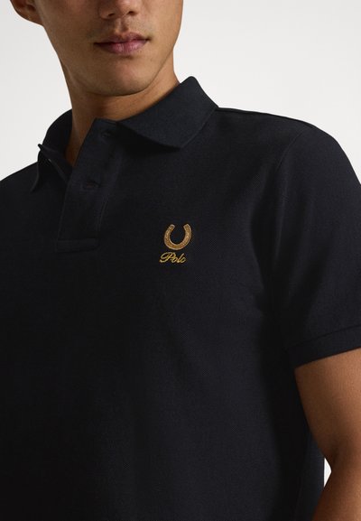 Jaunas vyras, dėvintis juodą polo marškinėlį su aukso siuvinėta arkliuko pasaga ir tekstu "Polo" ant krūtinės.