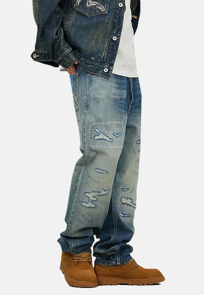 Jeans di denim blu con toppe scolorite e dettagli di cuciture visibili, abbinati a stivali marroni. Il design presenta una vestibilità comoda e un effetto usurato.