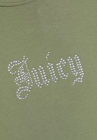 Πράσινο πουκάμισο από ύφασμα με τη λέξη "Juicy" γραμμένη με ασημί στρας σε διακοσμητική γραμματοσειρά.