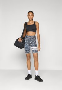 Black sports bra, patterned bike shorts in blue, black, and white. Model holds a black gym bag; wearing black sneakers and white socks.