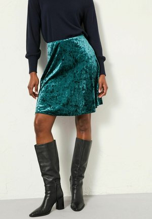 Türkisfarbener Samtrock mit einem ausgestellten Design, kombiniert mit knielangen schwarzen Lederstiefeln mit Blockabsatz und einem Reißverschluss an der Seite.