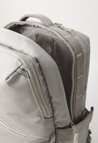 Pelēks mugursoma, kas izgatavota no izturīga neilona materiāla, ar vairākiem nodalījumiem, rāvējslēdzējiem un sānu kabatu; uz ārējās virsmas ir ievērojama režģa raksts.