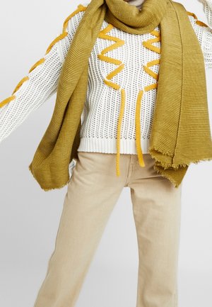 Person iført beige bukser, hvid strikket sweater med gule zigzag-detaljer og et stort sennepsgult tørklæde draperet omkring halsen.