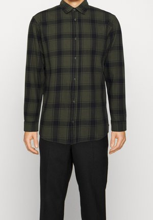 Mann trägt ein grün-schwarz kariertes, langärmliges Hemd mit Knöpfen und Kragen, kombiniert mit schwarzen Hosen, steht vor einem einfarbigen Hintergrund.