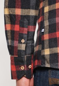 Flanelová košile s šedým, červeným a žlutým károvaným vzorem. Pozoruhodné detaily zahrnují dřevěné knoflíky a malou logo nášivku na boku.