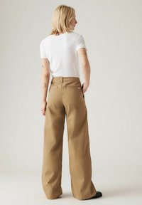 Een tan kleurige wijde broek van een stevige katoenmix, met achterzakken en een hoge taille. Gekoombineerd met een effen wit aansluitend t-shirt.
