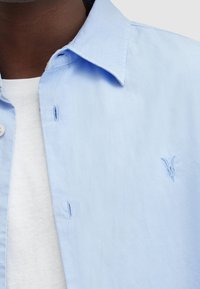 Camisetta azzurro chiaro con colletto a bottone, con un logo ricamato di testa di capra sul petto, indossata sopra una T-shirt bianca a girocollo.