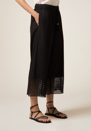 Kobieta w czarnych, szerokich, skróconych spodniach z siateczkową lamówką i czarnych, płaskich sandałach z ćwiekami i paskami, ręka w kieszeni.