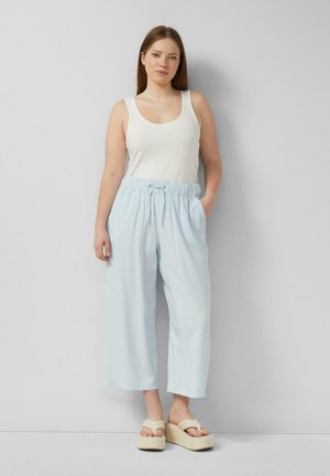 Femme debout contre un mur gris, portant un débardeur blanc, un pantalon large bleu clair à cordon, et des sandales plates-formes beige.