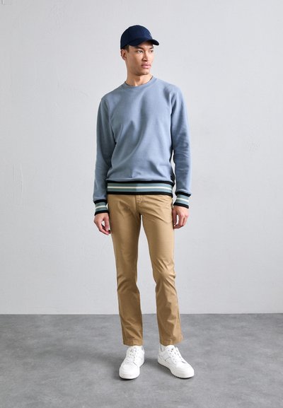 Light blue sweatshirt with striped black and teal cuffs, paired with tan chinos and white sneakers. Flat navy cap completes the outfit.