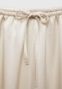 Bež hlače s elastikom u struku imaju glatku, satenastu teksturu, s vezicom za zatvaranje. Tkanina ima suptilni sjaj i minimalne šavove.