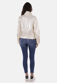 Giacca crema leggera e lucida con colletto alto, abbinata a jeans blu e sneakers bianche con accenti neri. Vista posteriore mostrata.