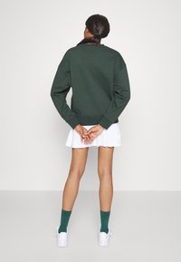 Sweat-shirt vert foncé à coupe décontractée, col rond et épaules tombantes, assorti à une jupe blanche plissée et des chaussettes vertes.