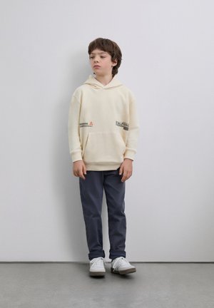 Sudadera con capucha color crema con bolsillo frontal y estampados gráficos. Combinada con pantalones azul marino y zapatillas blancas. Tela suave y corte relajado.