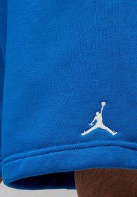 Niebieskie szorty z tkaniny z białym haftowanym logo przedstawiającym skaczącego koszykarza z piłką w pobliżu brzegu.