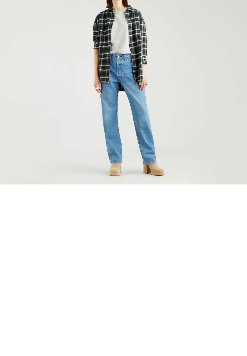 Femme portant des bottes compensées en daim beige, un jean large bleu clair, un t-shirt gris et une longue chemise à carreaux, debout devant un fond clair uni.
