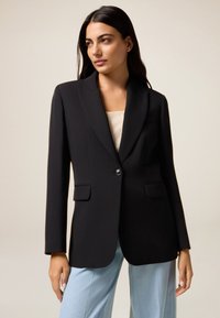 Blazer nero a un bottone con rever a punte, design minimalista, due tasche frontali e texture liscia. Abbinato a pantaloni azzurri chiari.