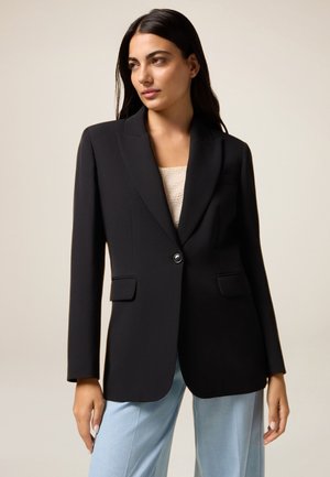 Blazer noir à un bouton avec un col cranté, design minimaliste, deux poches avant et texture lisse. Associé à un pantalon bleu clair.